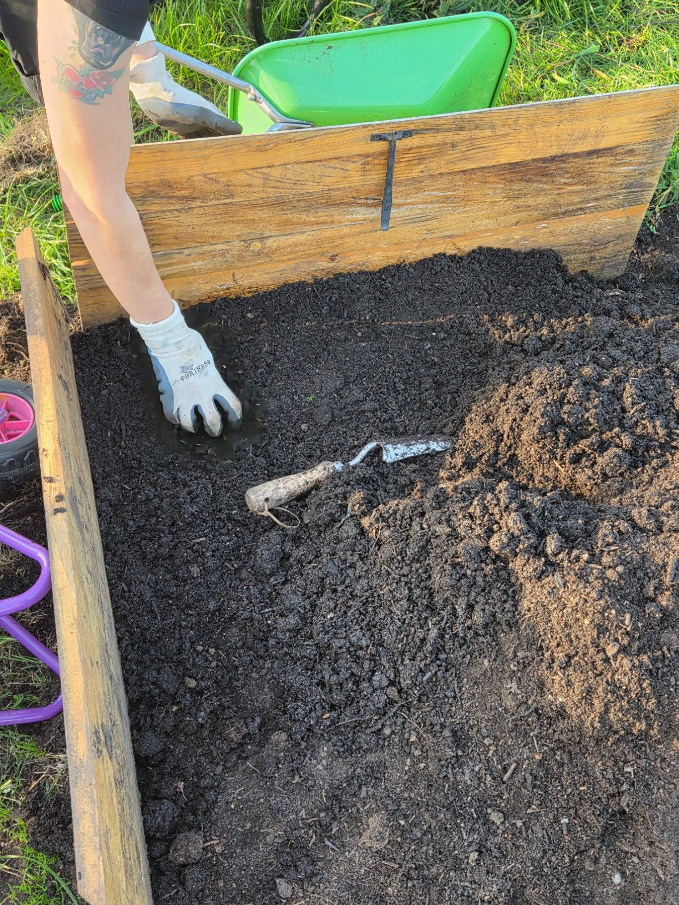 A hand in a glove digging in a garden bed with tools and soil visible.