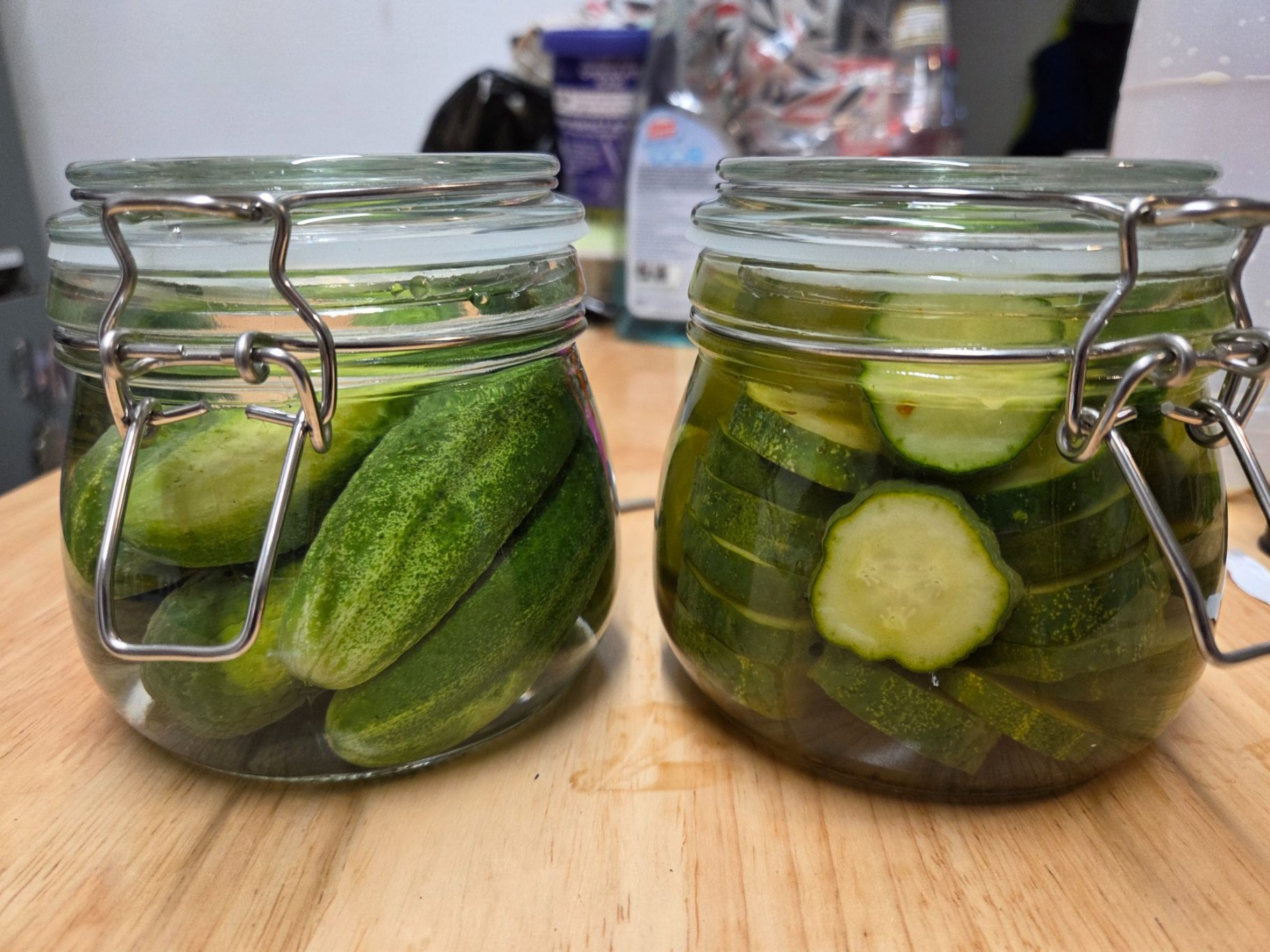 Two glass jars filled with cucumbers, one whole and one sliced, on a wooden surface.